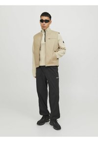 Beige mouwloos jasje over een crèmekleurige trui, gecombineerd met losse zwarte broek en zwarte schoenen. Heeft een ritszak en logo-accenten.