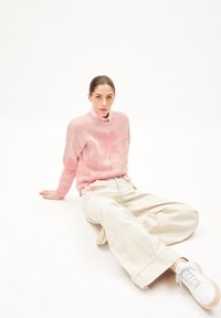 Rosa gestrickter Pullover mit Blumenmuster, beiger weiter Hose und weißen Sneakers. Model sitzt vor einem schlichten Hintergrund.