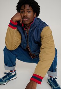Denim and beige bomber jacket with red and navy ribbed cuffs. Blue sneakers and rolled-up jeans with a checkered shirt visible underneath.