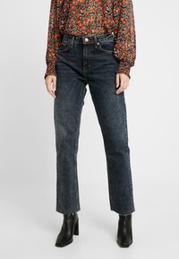 Jeans dritti blu scuro con vita alta, caratterizzati da un leggero effetto usurato e orlo a vivo. Abbinati a una blusa a fantasia floreale e stivaletti neri con tacco.