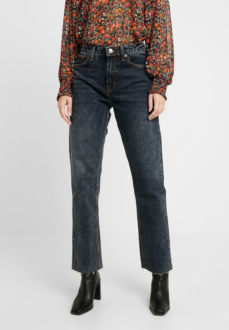 Jeans dritti blu scuro con vita alta, caratterizzati da un leggero effetto usurato e orlo a vivo. Abbinati a una blusa a fantasia floreale e stivaletti neri con tacco.