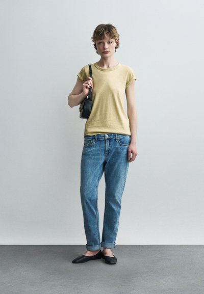 Νεαρό άτομο με κοντά μαλλιά που φοράει κίτρινο t-shirt, τζιν παντελόνι μπλε με γυρισμένα μπατζάκια, μαύρα φλατ παπούτσια και κουβαλάει μια μαύρη τσάντα ώμου.