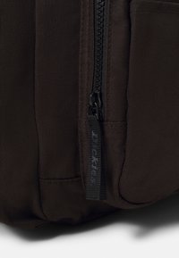 Bolsa de lona marrón vista lateral con un zipper negro y un tirador con la marca "Dickies". Incluye tejido texturizado con costuras visibles.