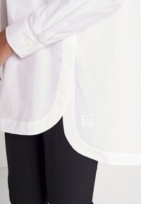 Hvid skjorte med knapper og buet kant samt lange ærmer, der har et subtilt logo trykt nær kanten. Sorte bukser ses også.