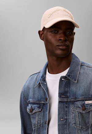 Chaqueta vaquera azul con dos bolsillos en el pecho, detalles de botones, camiseta blanca de cuello redondo y gorra beige clara con texto bordado.