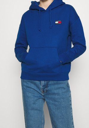 Osoba w niebieskiej bluzie z kapturem Tommy Jeans z przednią kieszenią i niebieskich dżinsach, stoi z rękami w kieszeniach bluzy na jednolitym tle.