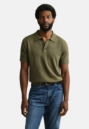 Man met baard die een olijfgroen gebreid polohemp met korte mouwen en een blauwe spijkerbroek draagt, staand tegen een eenvoudige lichte achtergrond.