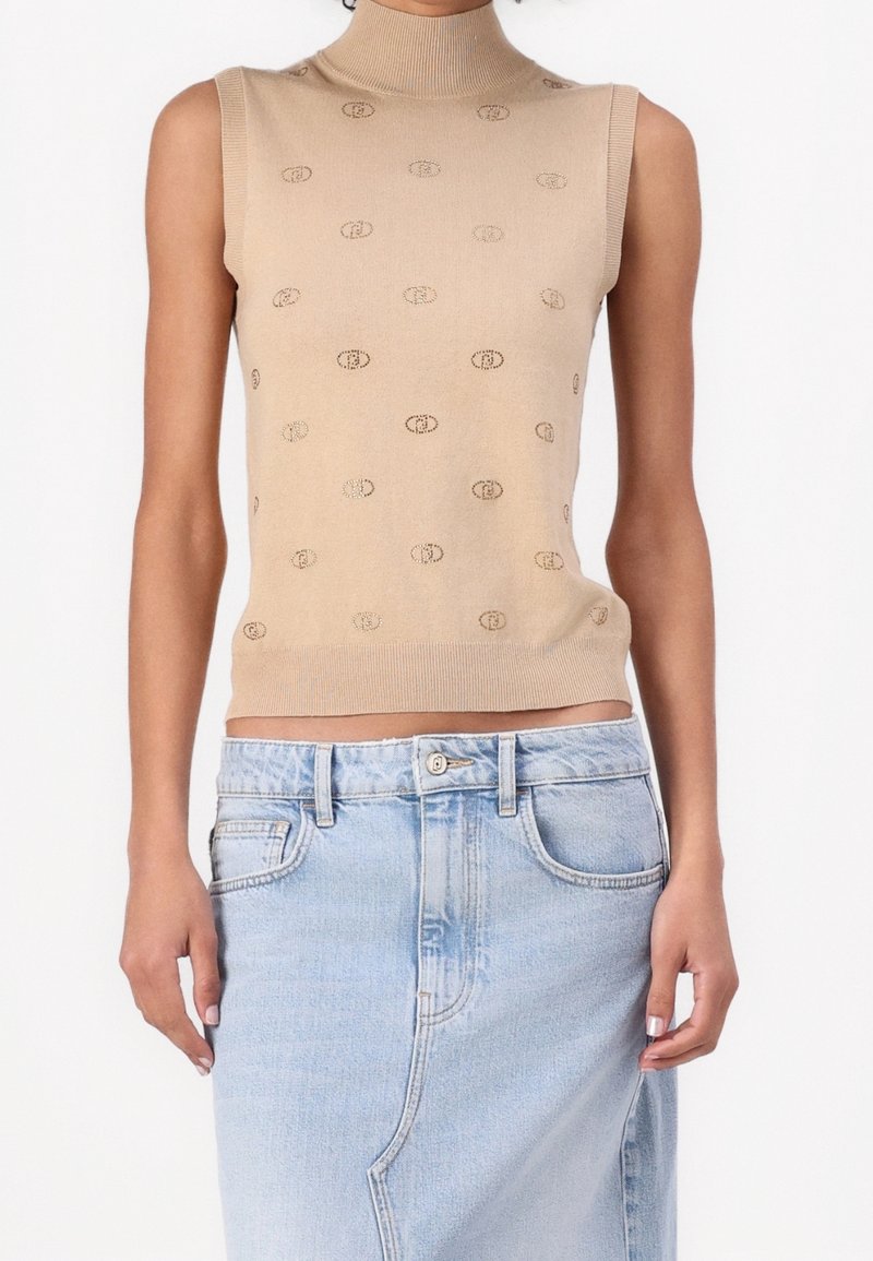 Pull sans manches beige à col roulé avec petits motifs ovales répétitifs, associé à une jupe en denim bleu clair taille haute.
