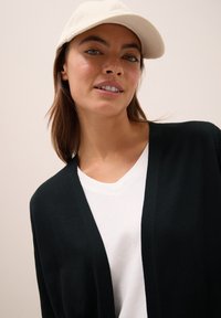 Zwarte gebreide cardigan met een diepe V-hals, gecombineerd met een witte T-shirt. Het model draagt een lichtgekleurde pet, gemaakt van een gestructureerde stof.