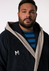 Mörkblå badrock med en beige huva och trim, tillverkad av mjuk frotté. Har en broderad "M" på vänster bröst.