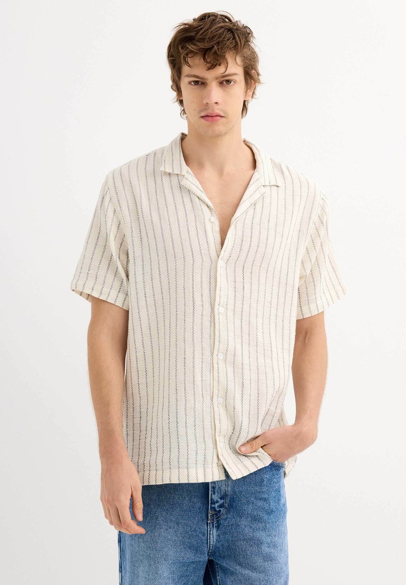 Camisa de manga corta, con cuello, en color crema con rayas verticales azules, que presenta un cierre de botones en la parte delantera y un corte relajado. Fabricada en tejido texturizado.
