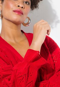 Roter gestrickter Pullover mit V-Ausschnitt, der ein strukturiertes Muster und Ballonärmel aufweist. Das Model trägt große silberne Creolen.