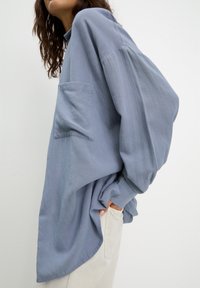 Chemise surdimensionnée bleu clair avec un col haut, des manches longues et une poche poitrine. Fabriquée en tissu doux, elle présente une silhouette fluide et décontractée.