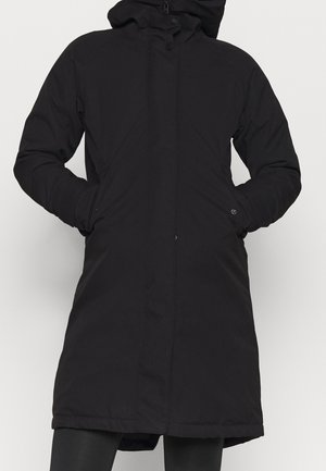 Abrigo de invierno negro con capucha, cremallera frontal y botones de presión, usado con las manos en los bolsillos laterales, cubriendo la parte superior de los muslos.