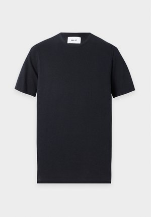 Jeune homme portant un t-shirt noir, un long short en jean foncé avec chaîne, des chaussettes noires et des mocassins bicolores, debout devant un fond uni.