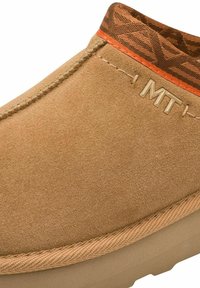 Chaussure slip-on en daim marron avec un dessus texturé à motifs marron, accent orange et détail en métal du logo "MT" sur le côté.