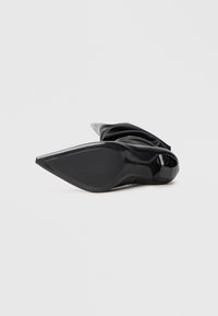 Zwarte puntige schoen van glad materiaal met een gevouwen ontwerp, voorzien van een slanke stilettohak en een gestructureerde zool voor grip.