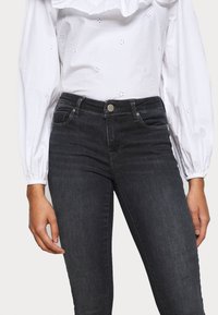 White shirt with textured eyelet design and ruffled neckline paired with dark gray slim-fit jeans featuring a button closure and front pockets.