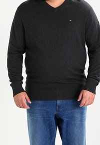 Mörkgrå v-stickad tröja med lång ärm, ribbade cuffs och nederkant. Materialet verkar mjukt, bärs över blåjeans. Litet logotypdetalj på bröstet.