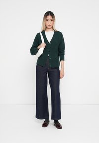 Cardigan tricoté vert avec un motif en losange, fermeture à boutons, porté sur une chemise blanche. Associé à un pantalon large en denim foncé et des chaussures noires.