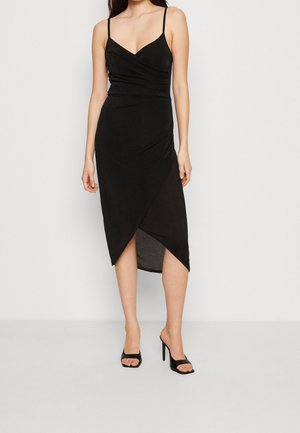 Femme portant une robe porte-feuille noire sans manches à fines bretelles et des chaussures noires à talons hauts et bouts ouverts, debout sur un fond blanc.
