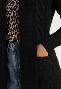 Czarny dzianinowy kardigan z wzorami warkoczowymi, ściągaczowymi mankietami i bocznymi kieszeniami, noszony na bluzce w panterkę i dżinsach.