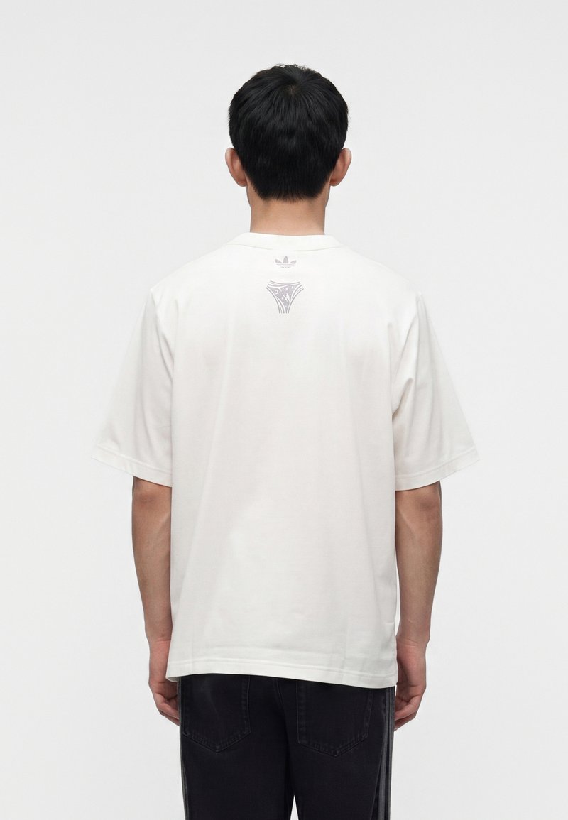 Uomo che indossa una t-shirt bianca a maniche corte con una piccola grafica e logo sulla parte superiore della schiena, abbinata a pantaloni neri.
