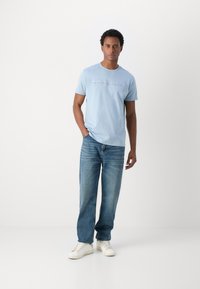 Lichtblauwe katoenen t-shirt met geborduurde tekst, gecombineerd met rechte blauwe denim jeans en witte sneakers. Eenvoudige, casual outfit.