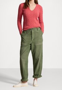 Czerwony sweter z dzianiny warkoczowej z dekoltem w serek i logo; oliwkowo-zielone, luźne spodnie cargo z kieszeniami; lekkie płócienne trampki.