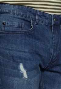 Calças de ganga em azul escuro com uma textura ligeiramente desgastada. Apresentam um bolso frontal, um pequeno rasgado na coxa e um fecho com botão.