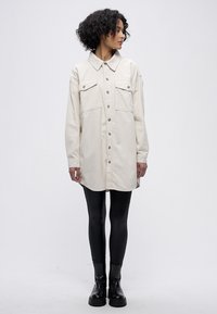 Robe chemise beige avec col à revers, boutonnage sur le devant et deux poches poitrine. Associée à des leggings noirs et des bottines. Tissu à la texture lisse.