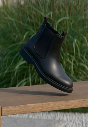 Bottines Chelsea en cuir noir avec des panneaux élastiques sur les côtés et des languettes, flottant au-dessus d'un banc en bois en extérieur avec un fond d'herbe.