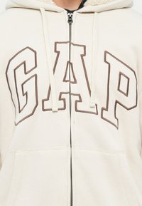 Sudadera con cremallera beige con grandes letras marrones contorneadas de GAP en el pecho y cordones blancos, mostrando la parte superior del torso.