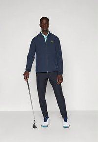 Jūras zila rāvējslēdzēja jaka, gaiši zils polo krekls zem tā, tumšas bikses un baltas golfa kurpes ar zilām detaļām, turot golfa nūju.