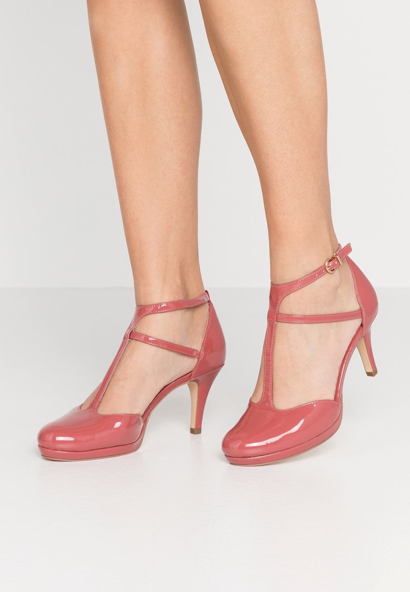Jambes portant des chaussures à talons hauts roses brillantes à bride en T avec boucle à la cheville, sur un sol blanc et un fond clair uni.
