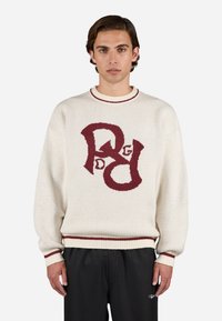 Creme strikket sweater med maroon logo-design på fronten, ribbet krave og manchetter, afslappet pasform og tekstureret stof. Sorte bukser båret.