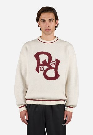 Cream knit sweater with maroon logo design on the front, ribbed collar and cuffs, relaxed fit, and textured fabric. Black trousers worn.