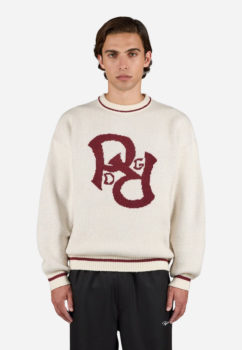 Cream knit sweater with maroon logo design on the front, ribbed collar and cuffs, relaxed fit, and textured fabric. Black trousers worn.