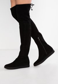 Bottes noires cuissardes en daim, dotées d'un design élégant, d'une forme structurée, d'une fermeture éclair intérieure et d'une semelle plate pour le confort.