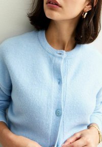 Cardigan tricoté bleu clair avec un col rond et une fermeture par un seul bouton. Sa texture douce et ses poignets côtelés ajoutent du détail.