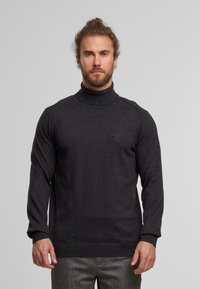 Mann mit Bart und gebundenem Haar, der einen dunkelgrauen Rollkragenpullover und graue kariierte Hosen trägt, steht vor einem schlichten Hintergrund.