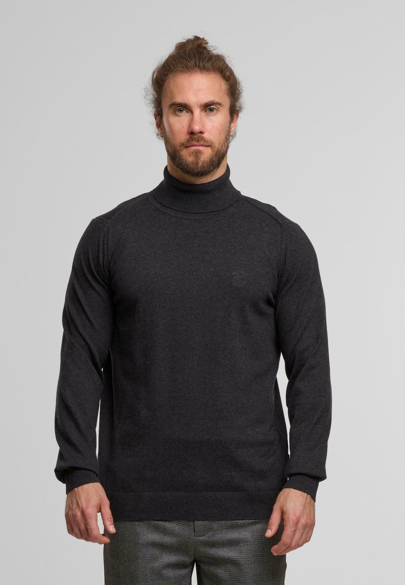 Mann mit Bart und gebundenem Haar, der einen dunkelgrauen Rollkragenpullover und graue kariierte Hosen trägt, steht vor einem schlichten Hintergrund.