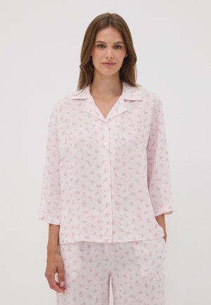 Mujer con pijama de color rosa claro con pequeño estampado floral, camisa abotonada con cuello y pantalones sueltos a juego.