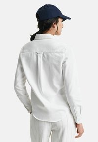 Weißes Langarmshirt mit Knopfmanschetten und abgerundetem Saum, kombiniert mit einer marineblauen Kappe. Der Stoff wirkt weich und strukturiert.