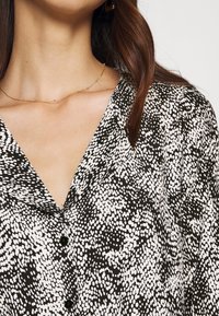 Sort-hvid mønstret bluse med V-udskæring, prydet med små polkaprikker og sorte knapper. Halskæden har tynde gyldne accenter.