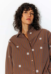 Chemise brune à boutons avec des rayures verticales bleues et de la broderie florale blanche. Elle comporte une poche sur le côté gauche et un col classique.