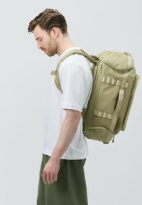 Khaki ryggsäck med flera fack, justerbara remmar och en slät, hållbar yta. Bärs på ena axeln av en person.