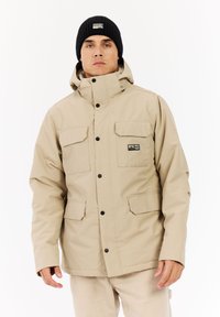 Beige vedenkestävä takki, jossa on korkea kaulus, kaksi rintataskua ja mustat napit. Yhdistetty mustan neuloksen pipoon.