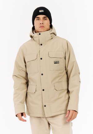 Beige wasserdichte Jacke mit hohem Kragen, zwei Brusttaschen und schwarzen Knöpfen. Getragen mit einer schwarzen Strickmütze.