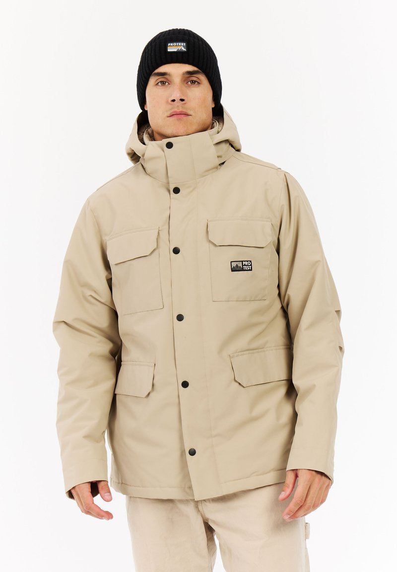 Beige vedenkestävä takki, jossa on korkea kaulus, kaksi rintataskua ja mustat napit. Yhdistetty mustan neuloksen pipoon.